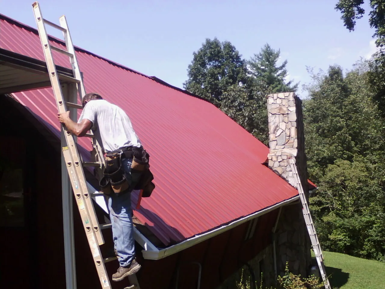 Roofer on a ladder beside a red metal roof and stone chimney — Soffit & Fascia Repair by Roofing Brothers in Munroe Falls